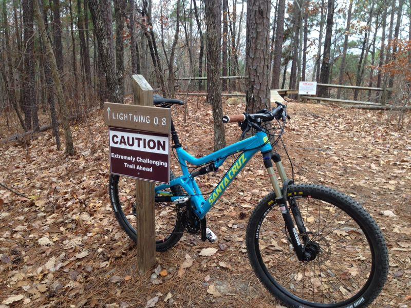A mountain bike leaning against a wooden sign in a wooded area. The sign reads "Caution: Extremely Challenging Trail Ahead" and is labeled "Lightning 0.8." The ground is covered with fallen leaves, and the trees are sparse, suggesting a trail setting. Coldwater Mountain mountain bike trail.