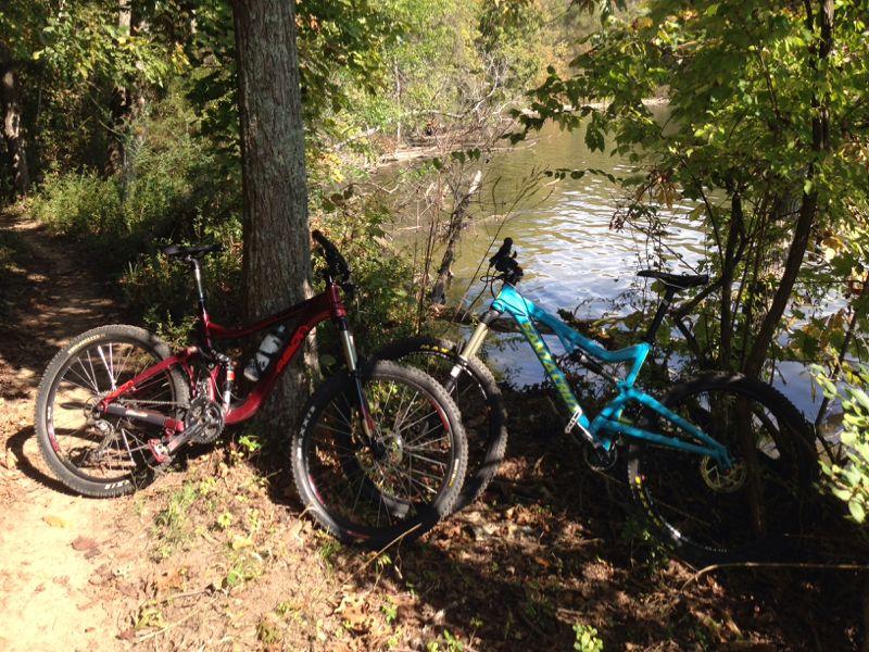 Two mountain bikes, one red and one turquoise, are leaning against a tree near a riverbank, surrounded by lush greenery and dappled sunlight filtering through the leaves. Lock 4 mountain bike trail.