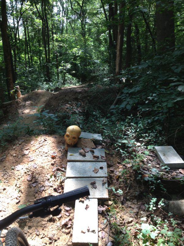 A narrow pathway through a wooded area, featuring a yellow skull placed on a stack of flat stones. The path is surrounded by lush greenery, and there are scattered leaves on the ground. A bike handlebar is partially visible in the foreground. In the background, a dirt jump can be seen. Back Yard Trails mountain bike trail.