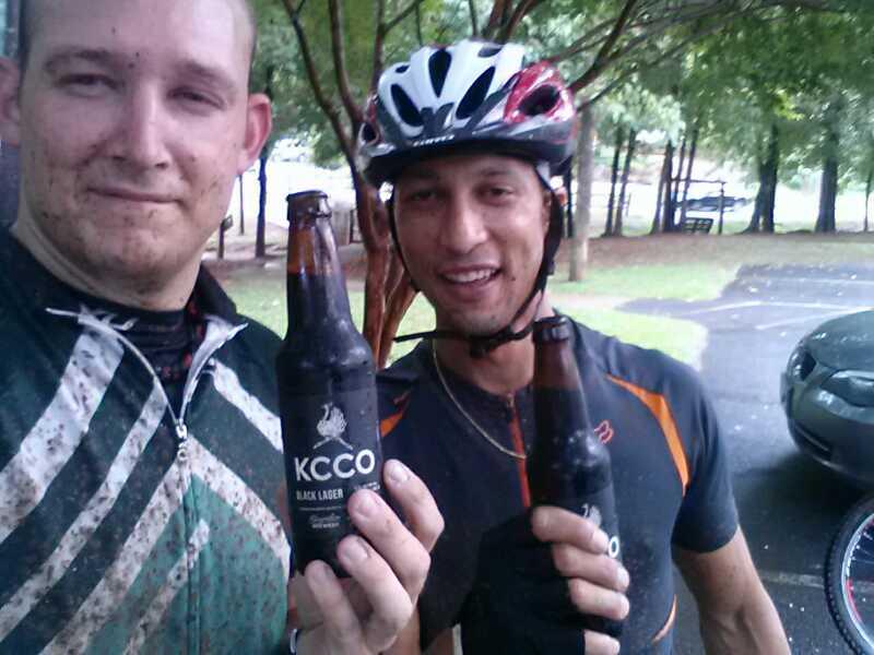 Two cyclists stand outdoors, both holding bottles of KCCO Black Lager. They are wearing cycling gear, with visible dirt on their clothing, suggesting they have just finished a ride. One cyclist wears a white helmet and has a slight smirk, while the other is smiling broadly. In the background, trees and a parked car are visible. Taylor Randahl Memorial Mountain Bike Trails At Olde Rope Mill Park mountain bike trail.