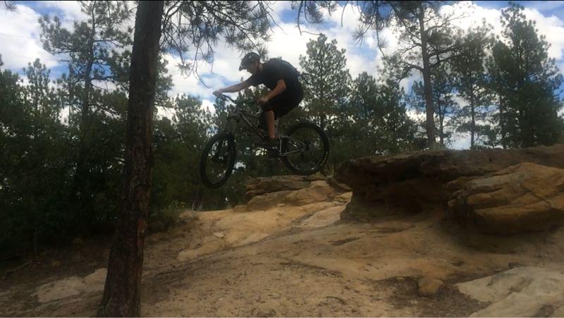 A mountain biker performing a jump off a rocky terrain in a forested area, with trees and a blue sky in the background. Palmer Park mountain bike trail.