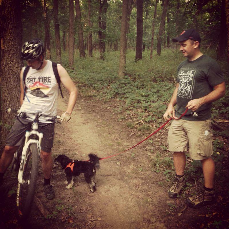 Two men in a wooded area, one wearing a tank top and biking shorts with a helmet, looking at his mountain bike. The other man, in a t-shirt and shorts, is holding a red leash attached to a small dog wearing an orange harness. The scene captures a casual outdoor moment on a hiking or biking trail. Swope Park Trail mountain bike trail.