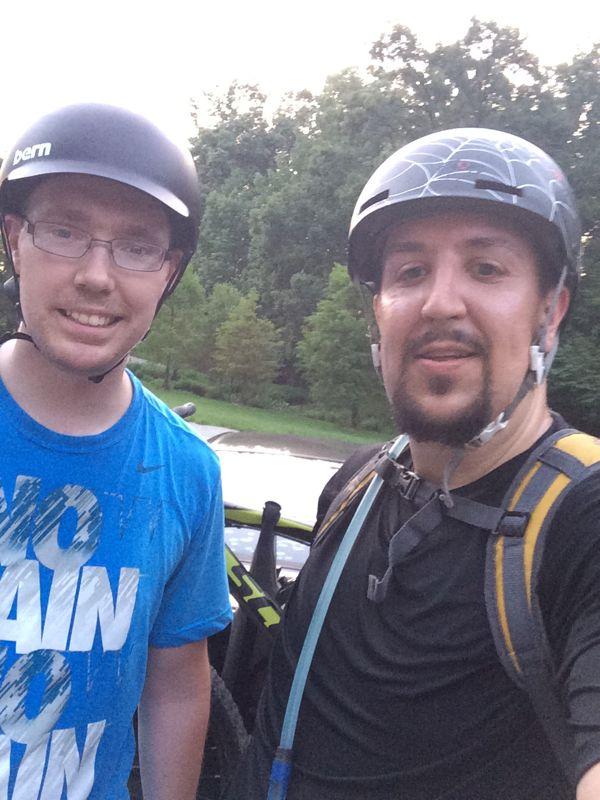 Two individuals wearing helmets stand together for a selfie outdoors. One person is wearing a blue shirt with the words "NO PAIN NO GAIN," while the other is dressed in a dark shirt and has a backpack. In the background, there are trees and a glimpse of a vehicle. The scene suggests they have been engaged in an outdoor activity, likely biking. Waverly Park mountain bike trail.