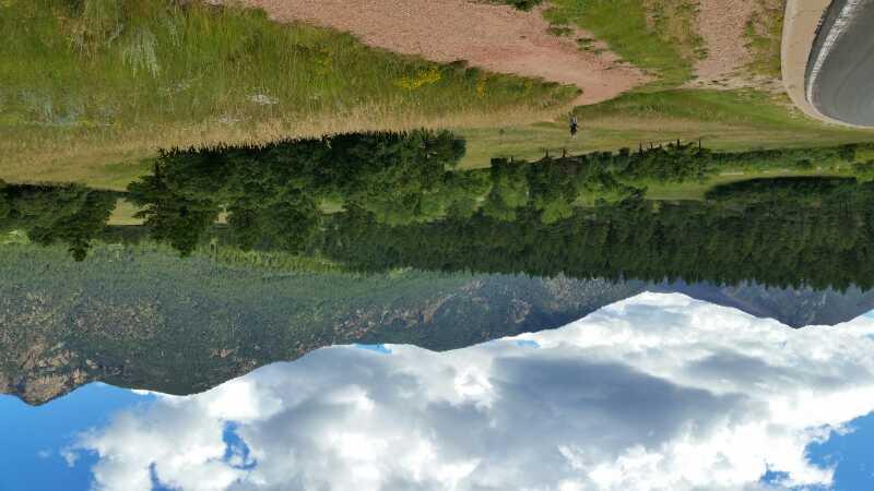 Scenic landscape featuring lush green fields and trees, with mountains in the background under a partially cloudy sky. A dirt path runs along the edge of the image, suggesting a natural outdoor area suitable for hiking or exploration. Falcon Trail mountain bike trail.