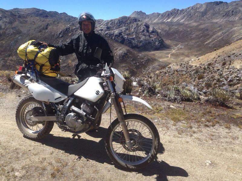 A person in a black jacket and helmet stands next to a white motorcycle parked on a dirt road. The motorcycle has a yellow backpack secured on the back. In the background, there are rugged mountains and a clear blue sky. The scene depicts a remote outdoor landscape, suggesting an adventure or travel experience. Valle De MifafÃ­ mountain bike trail.