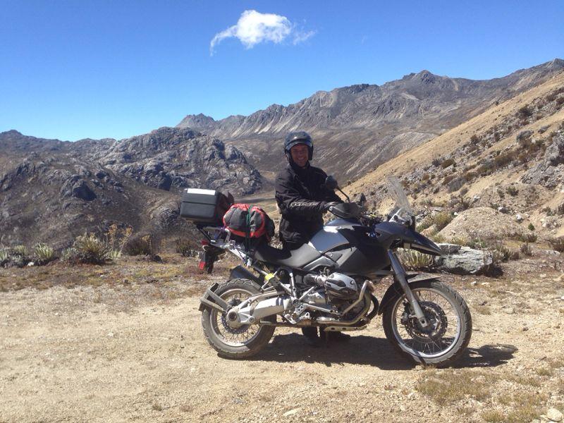 A motorcyclist wearing a helmet and riding gear poses next to a black motorcycle on a rugged, mountainous landscape. The background features rocky hills and a clear blue sky with a few clouds, suggesting an outdoor adventure. The motorcycle is equipped with luggage, indicating that the rider is likely on a long journey. Valle De MifafÃ­ mountain bike trail.