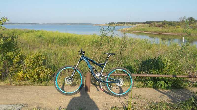 A mountain bike with blue accents parked on a sandy path beside a tranquil lake, surrounded by lush green vegetation and a clear sky in the background. Northshore Trail mountain bike trail.