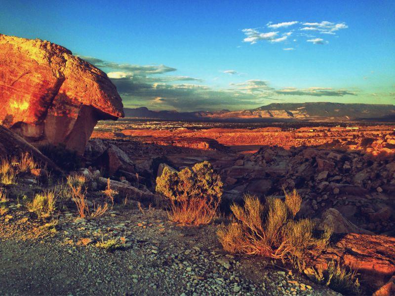 Scenic view of a rocky landscape at sunset, featuring vibrant orange and purple hues in the sky, a large boulder in the foreground, and distant mountains under a partly cloudy sky. The terrain is dotted with sparse vegetation, creating a serene and natural atmosphere. Lunch Loops mountain bike trail.