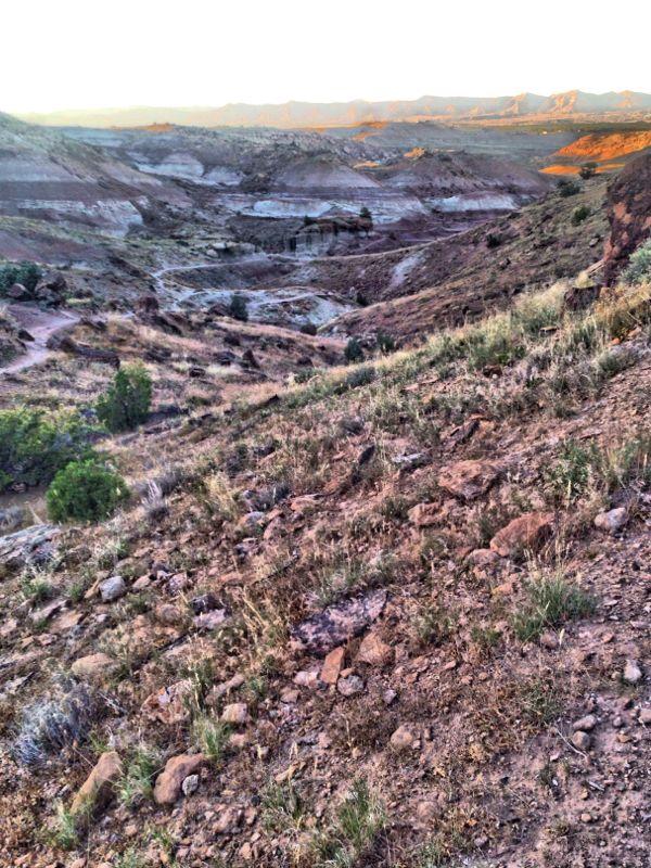 A scenic view of a rocky, arid landscape featuring rolling hills and sparse vegetation, with layers of earth exposed in shades of brown and gray. The soft sunlight illuminates the terrain, casting long shadows and highlighting the natural textures of the ground. In the distance, more rugged hills and mountains can be seen against a clear sky. Pet - A - Kes mountain bike trail.