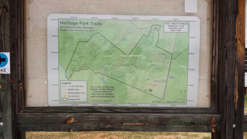 A map displaying the trails of Heritage Park in Oconee County, Georgia. The map includes various marked trails, including the Long Lake Trail, Westside Trail, and Heritage Park Trail, along with a legend to identify the trails. The background features a light green topographic representation of the park's terrain, with text indicating trail distances and a note to refer to the county's website for more information. The map is mounted on a wooden structure, surrounded by natural scenery. Heritage Park mountain bike trail.