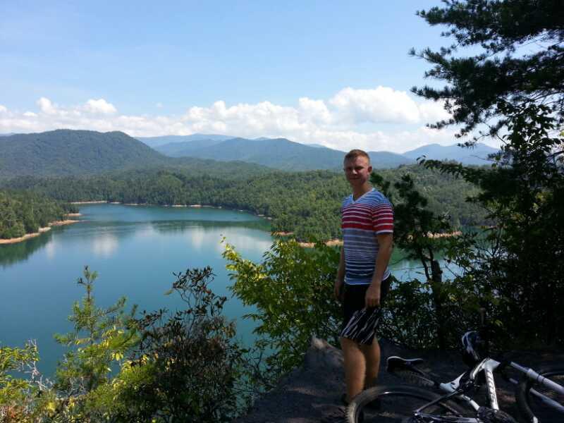 A person standing on a rocky overlook with a scenic view of a calm lake surrounded by lush green forests and mountains under a blue sky with fluffy white clouds. A bicycle is resting nearby on the ground. Tsali Left Loop mountain bike trail.