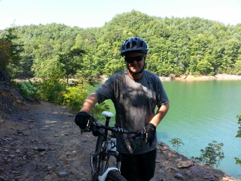 A mountain biker poses next to his bike on a rocky trail overlooking a serene lake, surrounded by lush green hills and trees on a sunny day. Tsali Left Loop mountain bike trail.