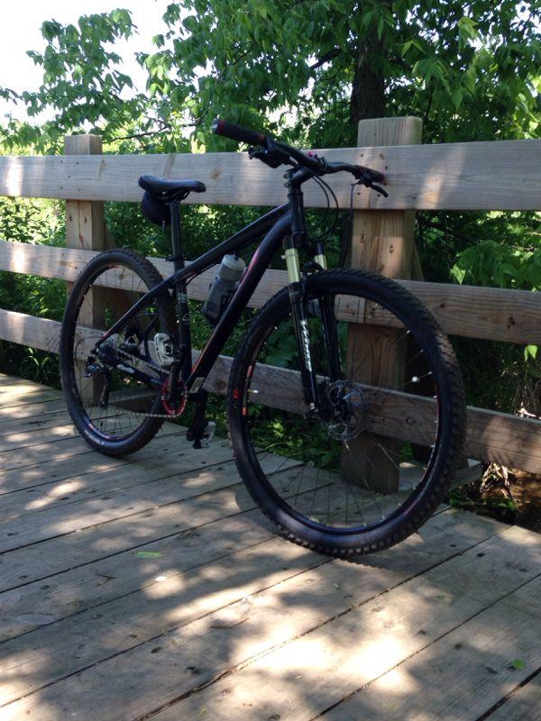 A black mountain bike parked on a wooden bridge surrounded by greenery. The bike has a water bottle holder and features visible suspension components, with sunlight filtering through the trees in the background. Burchfield mountain bike trail.