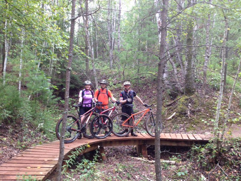 Three mountain bikers pose on a wooden bridge in a forested area, surrounded by tall trees and greenery. They are wearing helmets and cycling gear, each holding their mountain bikes, while smiling at the camera. Cuyuna Lakes mountain bike trail.