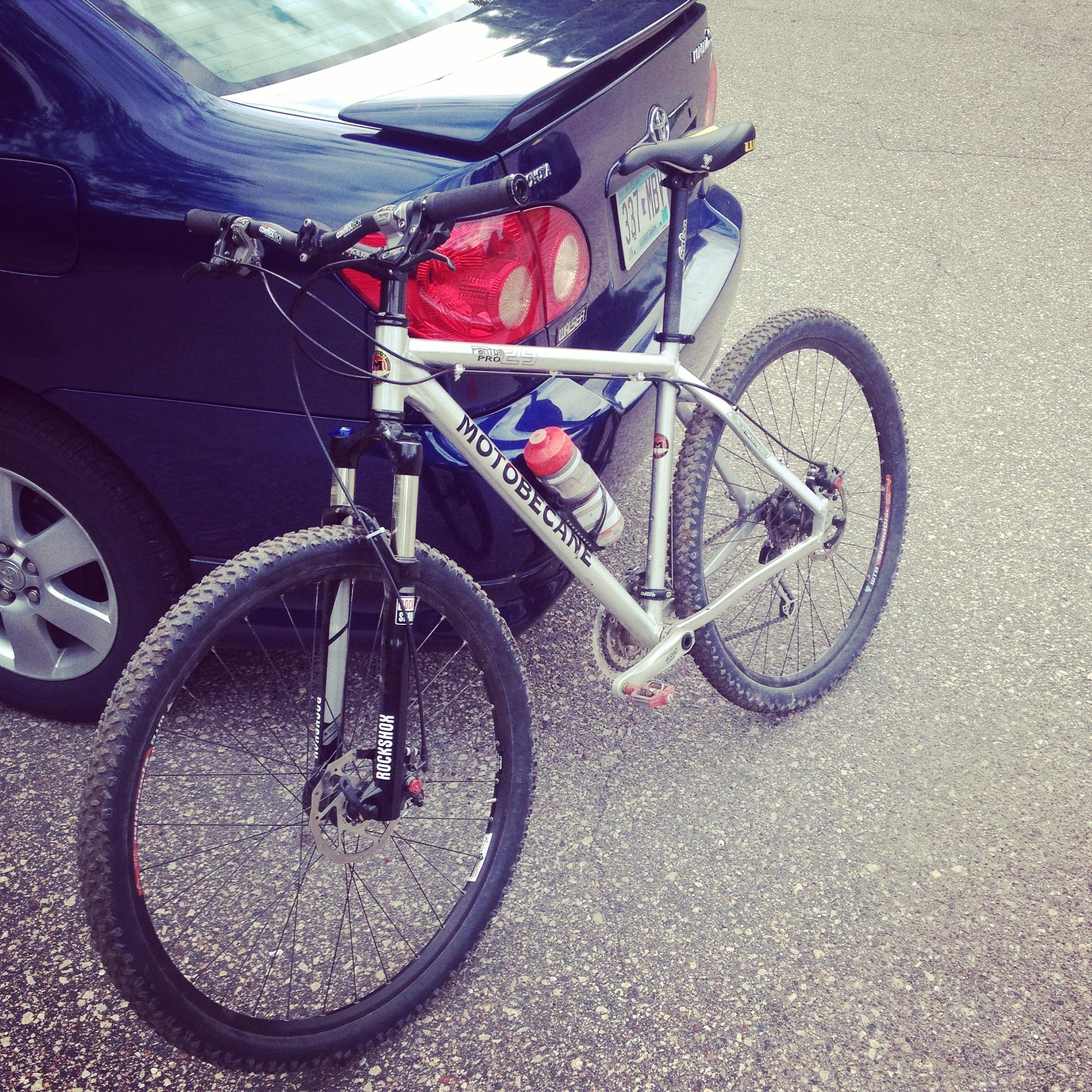 Motobecane Fantom 29 FS: A silver mountain bike with a water bottle attached, parked next to a blue car on a gravel road. The bike features thick tires and a front suspension.
