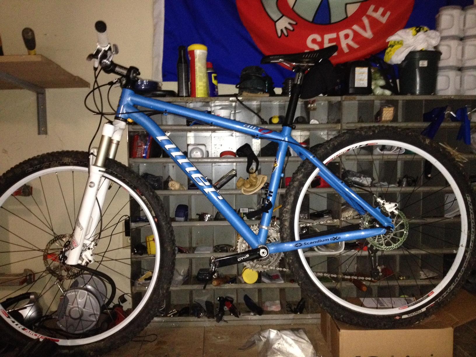 Niner Air 9: Mountain bike leaning against a storage shelf filled with various items. The bike features a blue frame, white front fork, and has dirt on the tires, indicating recent use. A red flag with a logo is visible in the background, along with containers and tools on the shelf.