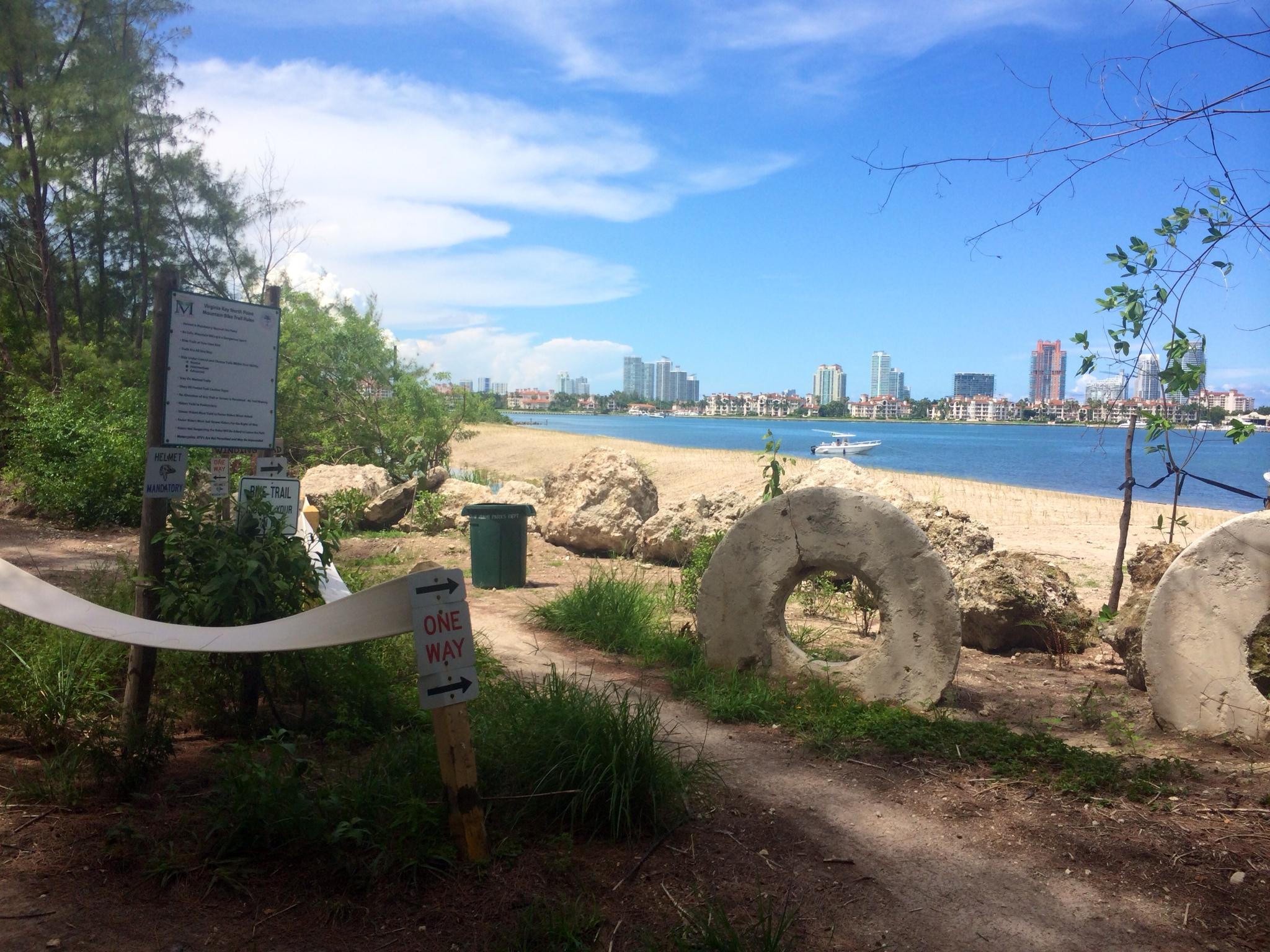 A scenic view of a beach pathway leading to the water, with lush greenery on either side. In the foreground, a signpost indicates trail directions, along with a trash bin nearby. The beach features fine sand, and a boat is visible in the water, with city buildings in the background under a bright blue sky with scattered clouds. Virginia Key North Point mountain bike trail.