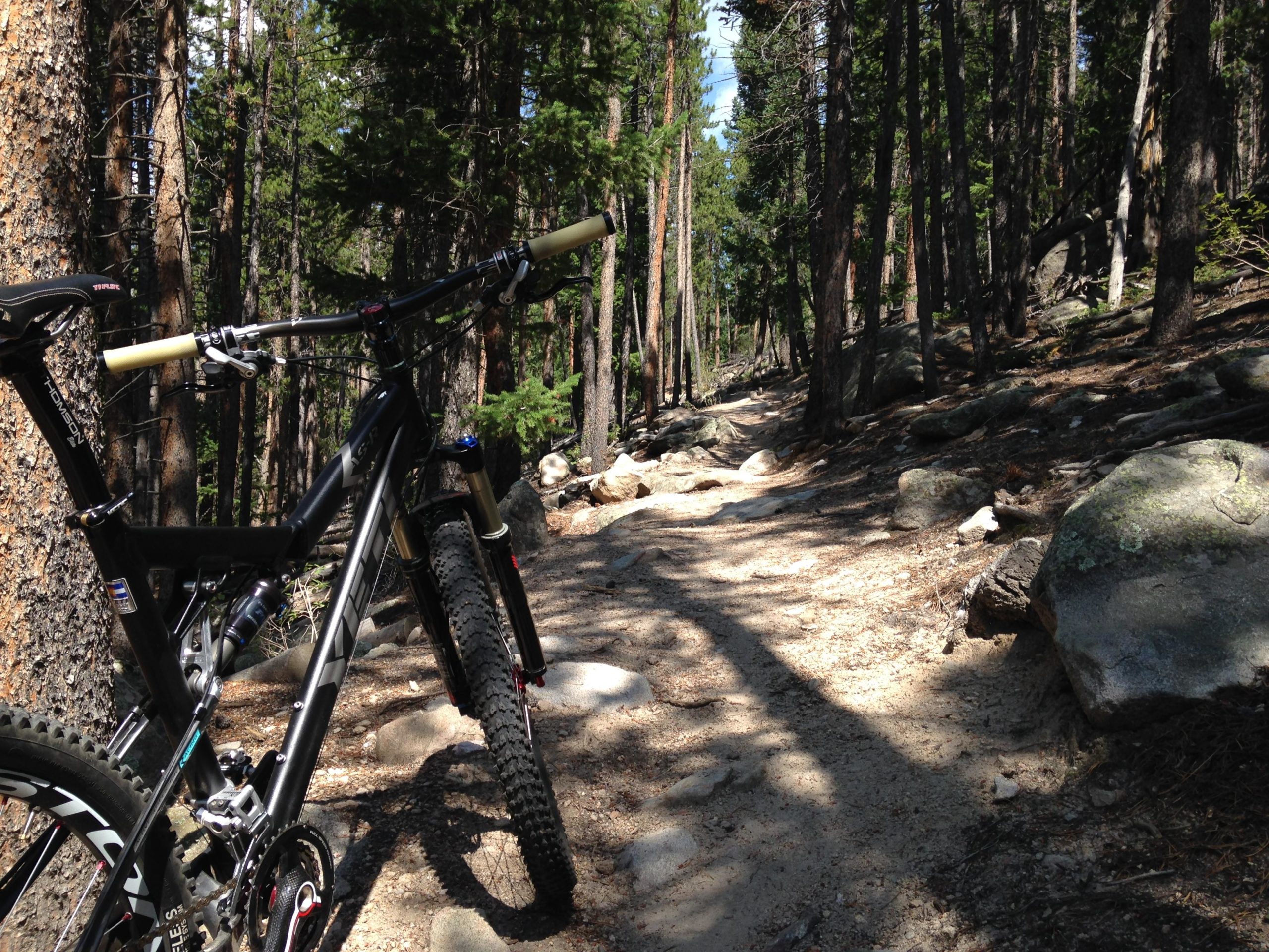 A mountain bike resting next to a rocky trail surrounded by tall trees and dappled sunlight in a forested area. Golden Gate Canyon State Park mountain bike trail.