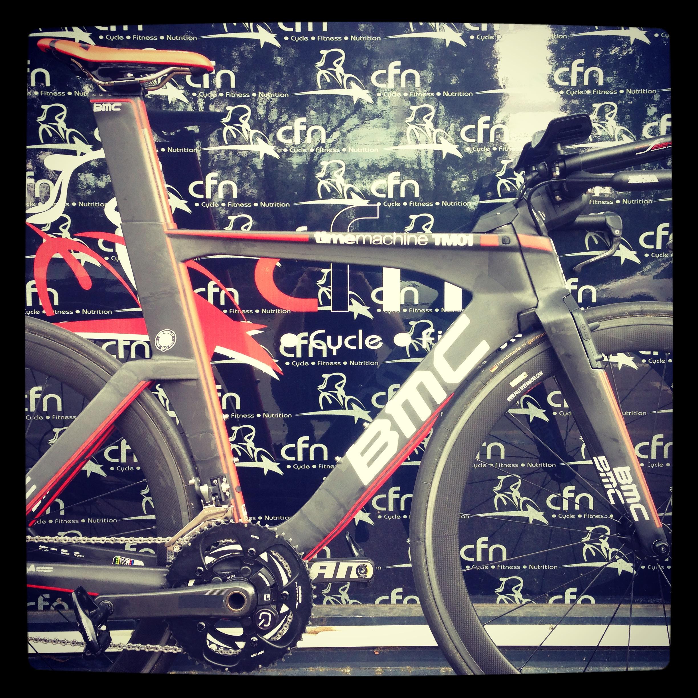 Alt text: A close-up view of a BMC Time Machine TM01 triathlon bike, featuring a sleek design with a gray and red frame, aerodynamic wheels, and a detailed gear system. The background displays a cycling-themed logo.