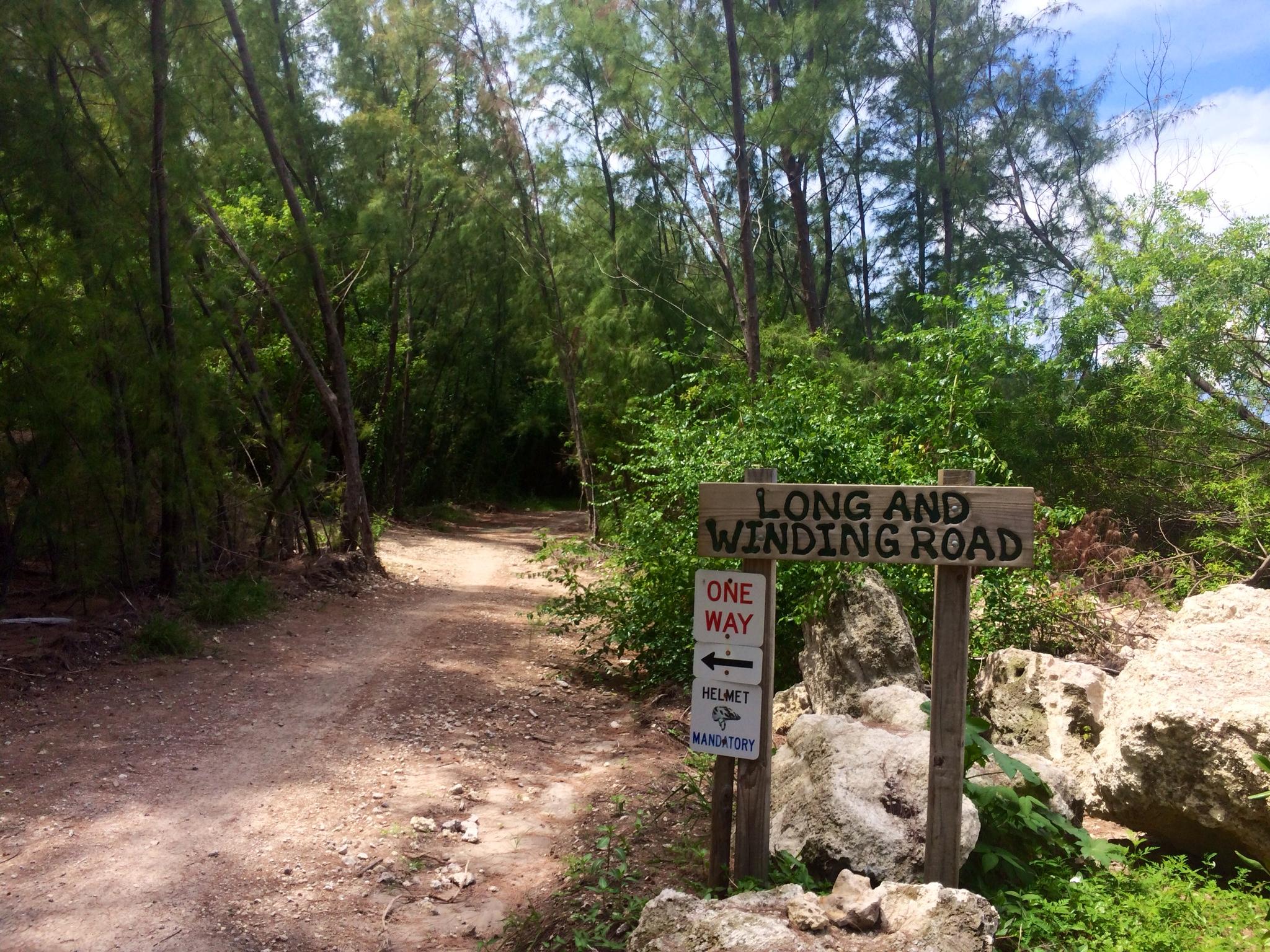 A dirt path winding through a lush green forest, with a wooden sign that reads "LONG AND WINDING ROAD" and additional signage indicating "ONE WAY" and "HELMET MANDATORY." The scene is bright and sunny, with tall trees lining the path and rocky outcrops in the foreground. Virginia Key North Point mountain bike trail.