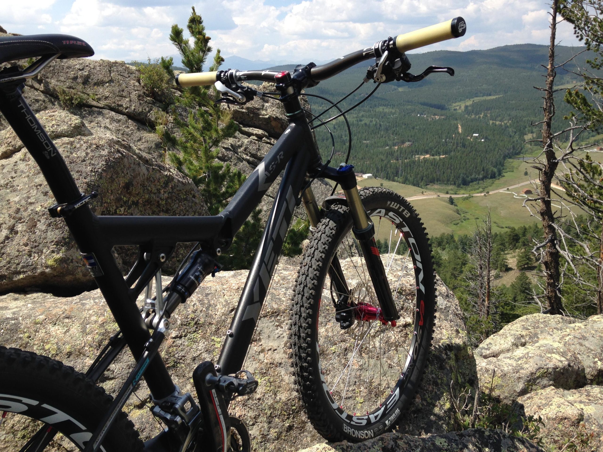 A black mountain bike is positioned on a rocky outcrop, overlooking a lush green valley and hills in the background. The bike features prominent suspension mechanics, thick tires, and handles with grips. The sky is partly cloudy, and there are scattered pine trees in the scene. Golden Gate Canyon State Park mountain bike trail.