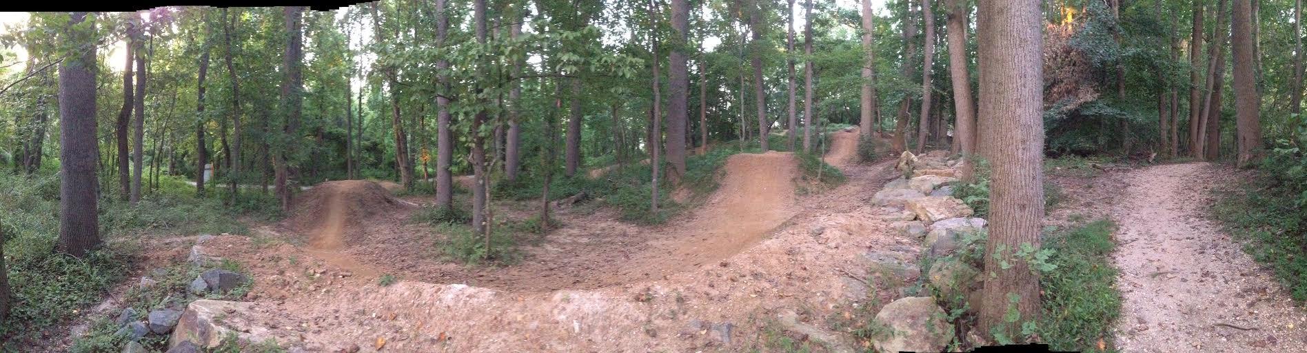A panoramic view of a wooded area featuring dirt bike trails and jumps, surrounded by tall trees and lush greenery. The image showcases a natural landscape with a mixture of dirt paths and rocky features, indicating an off-road biking or hiking trail. Rockburn Branch Park mountain bike trail.