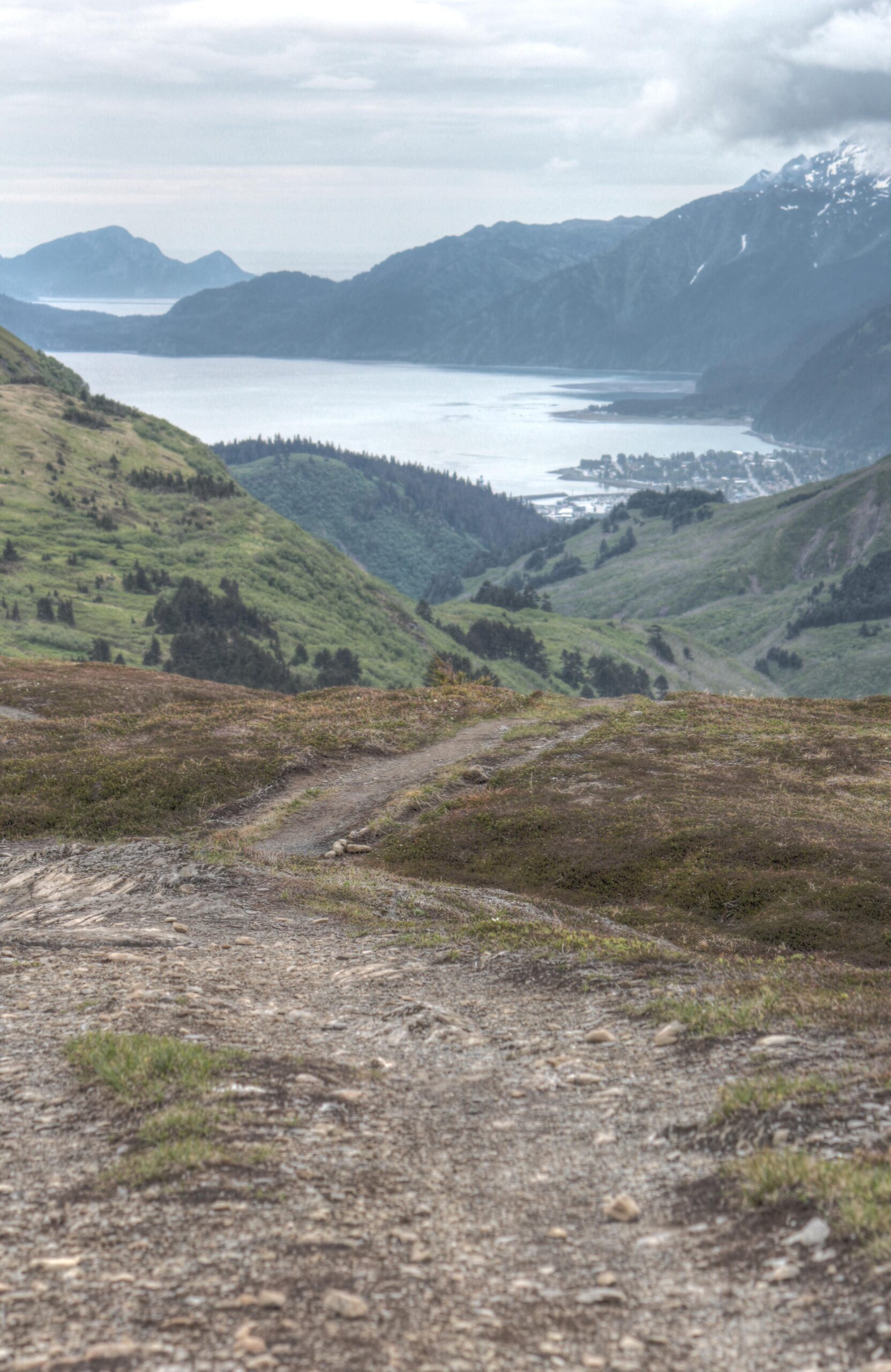 A scenic view from a mountain trail overlooking a lush green valley and a coastal town near the water. The landscape includes rolling hills, a winding dirt path leading downward, and distant mountains under a cloudy sky. Lost Lake Trail mountain bike trail.