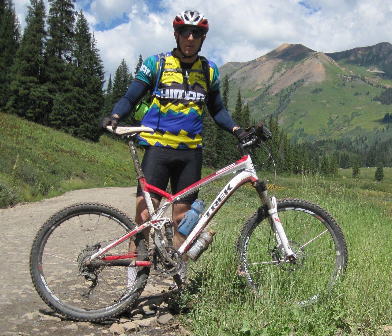 Trek Top Fuel 8: A mountain biker in a colorful jersey stands beside a red and white bike on a gravel path, surrounded by green grass and tall trees. Majestic mountains are visible in the background under a partly cloudy sky. The biker wears a helmet and sunglasses, with a water bottle attached to the bike.