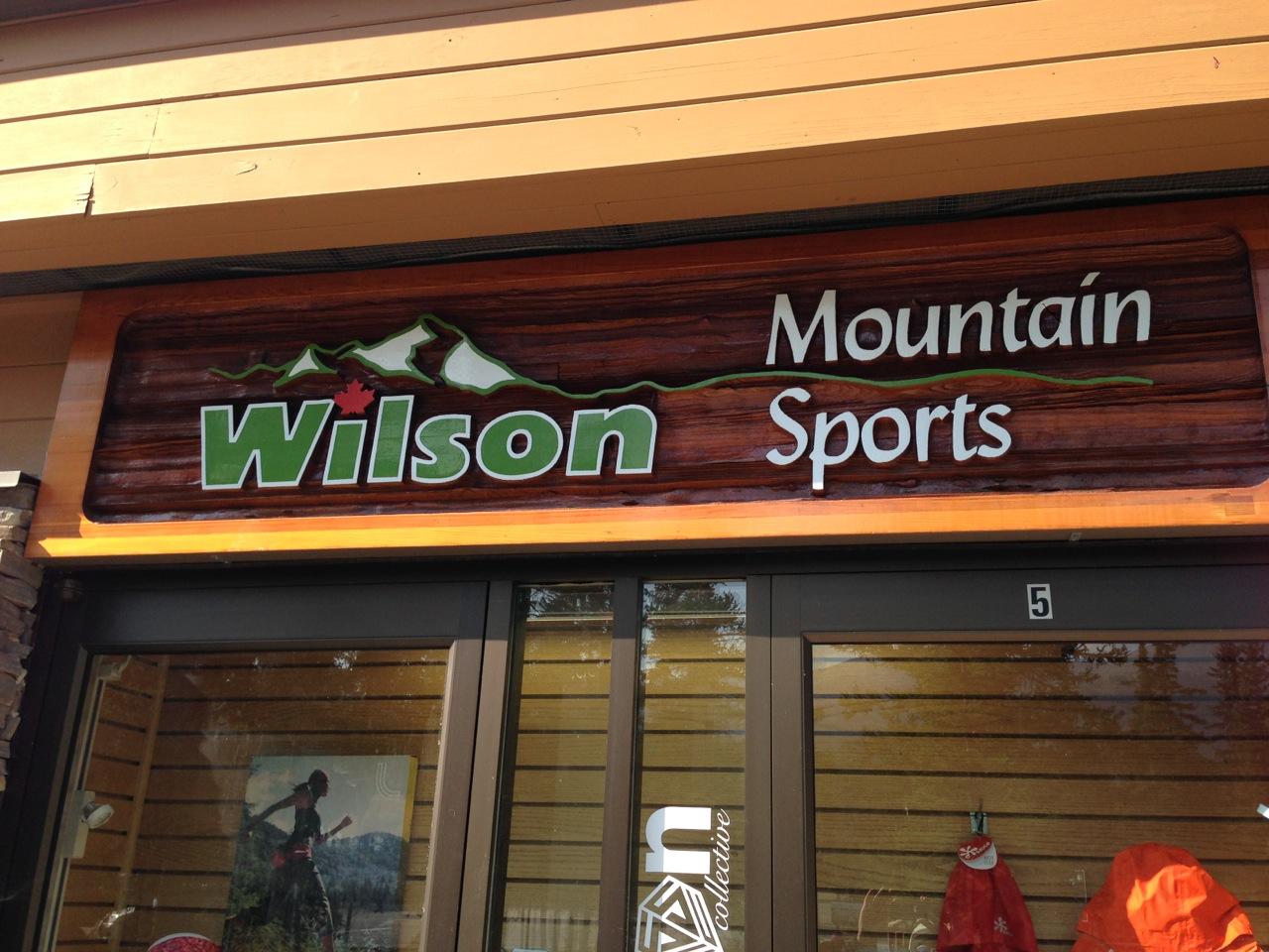 Signage for "Wilson Mountain Sports" featuring a wooden background and mountain graphics, displayed above the entrance to the store. The sign includes the name "Wilson" in large green letters and "Mountain Sports" in white letters.