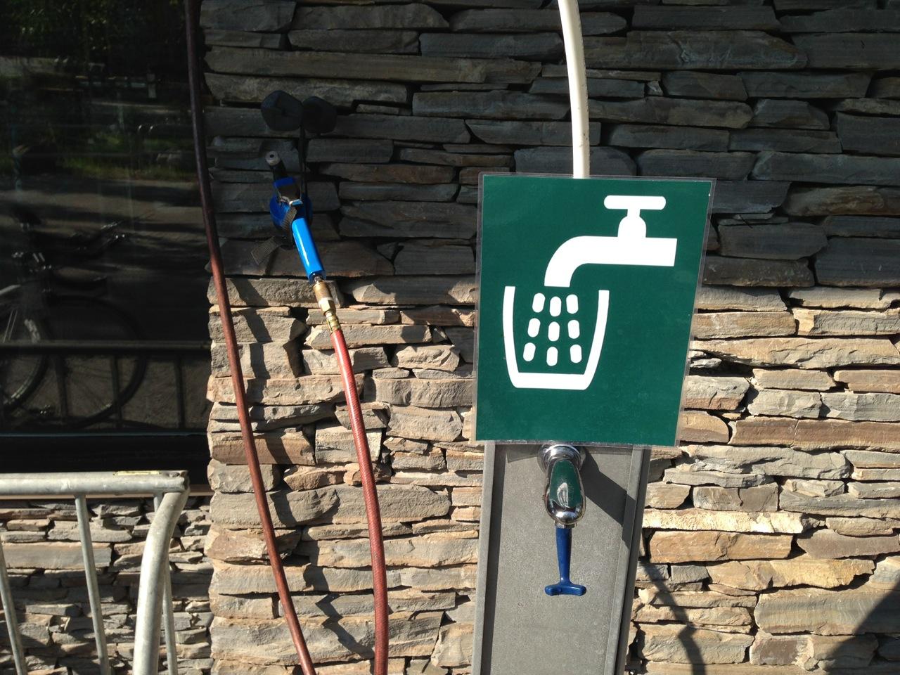 A wall-mounted water tap with a green sign displaying a faucet icon and water droplets, set against a textured stone background. A blue hose and a red hose are attached nearby.