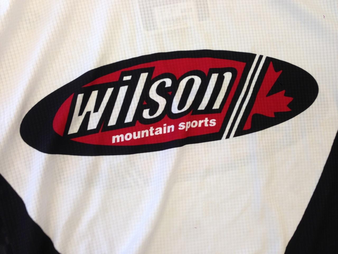 Image of a sports shirt featuring the Wilson logo. The logo is prominently displayed in black and red colors, with the text "wilson" and "mountain sports" underneath. A maple leaf design is incorporated into the logo. The background of the shirt is white with a textured pattern.
