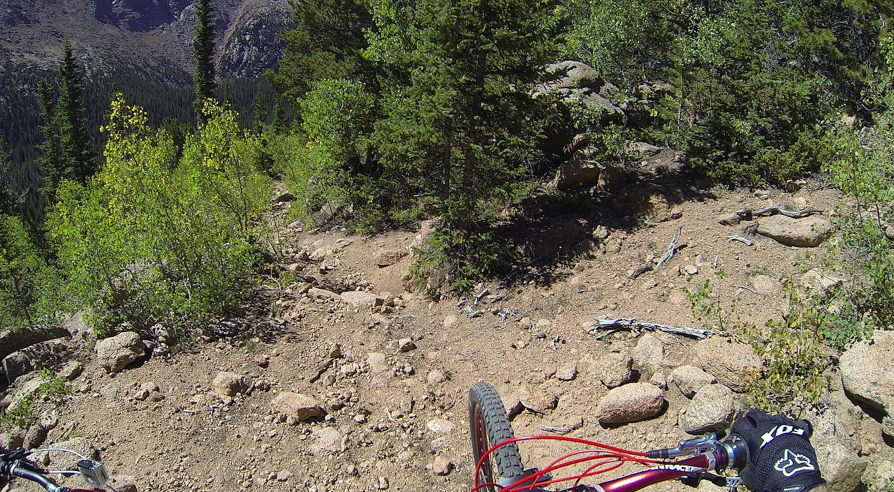 A cyclist's perspective on a rocky mountain biking trail, surrounded by trees and mountainous terrain. The view captures the bike's handlebars and front wheel, emphasizing the rugged path ahead, which is lined with rocks and dirt. Elk Park Trail mountain bike trail.