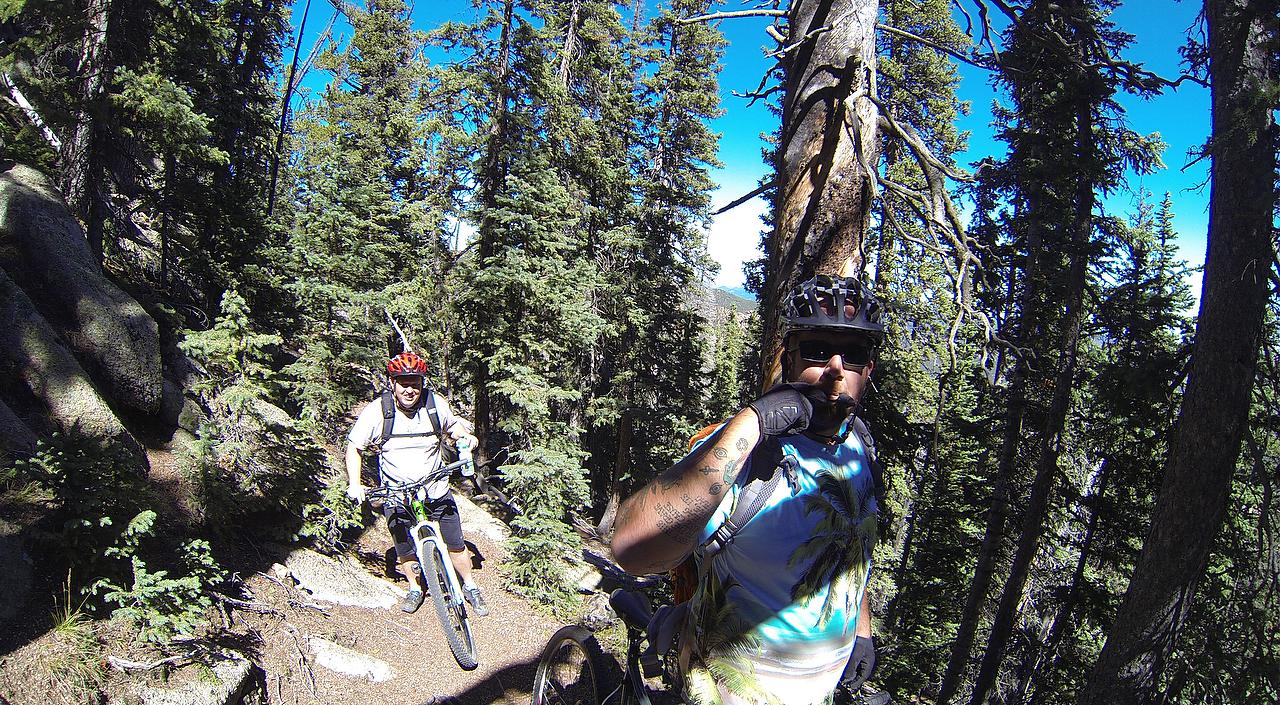 Two mountain bikers are riding along a narrow trail surrounded by dense forest. One biker, wearing a helmet and sunglasses, is playfully posing while holding up a finger to his lips, signaling for quiet. The other biker, also wearing a helmet, has paused to adjust his bike. Tall pine trees and rocky terrain make up the background under a clear blue sky. Elk Park Trail mountain bike trail.