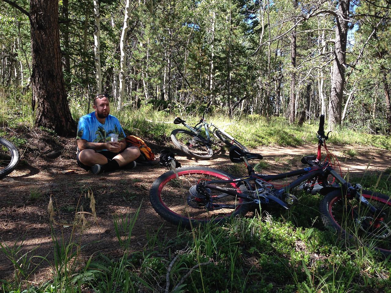 A person sitting cross-legged on the ground in a forested area, wearing a colorful t-shirt, holding a drink. Mountain bikes are resting on the ground nearby, surrounded by tall grass and trees. Longs Ranch Road mountain bike trail.
