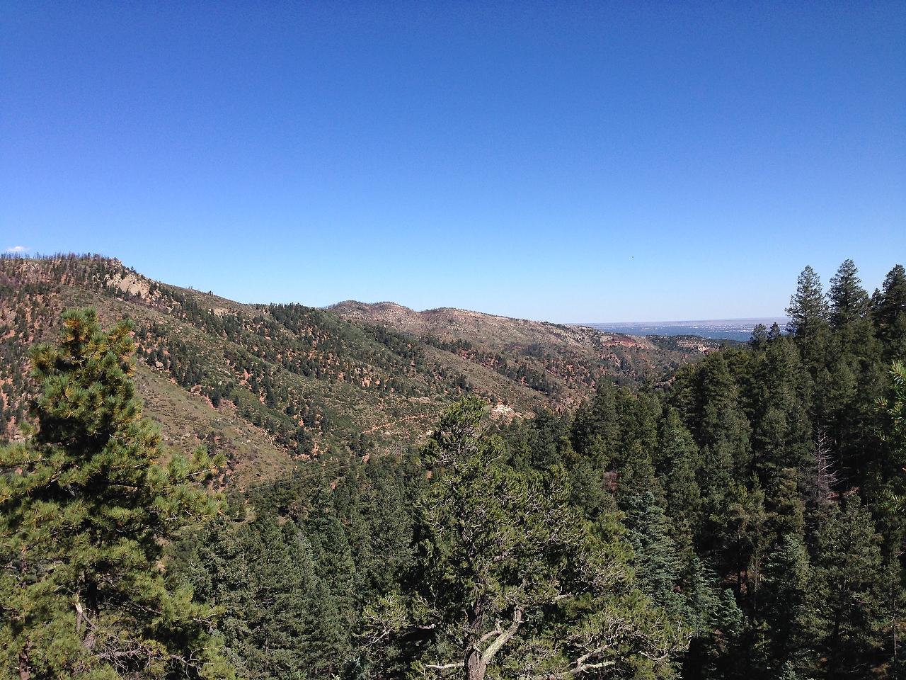 A panoramic view of rolling hills and densely wooded mountains under a clear blue sky. The landscape features a mix of evergreen trees and rocky outcrops, with a distant horizon visible in the background. Longs Ranch Road mountain bike trail.
