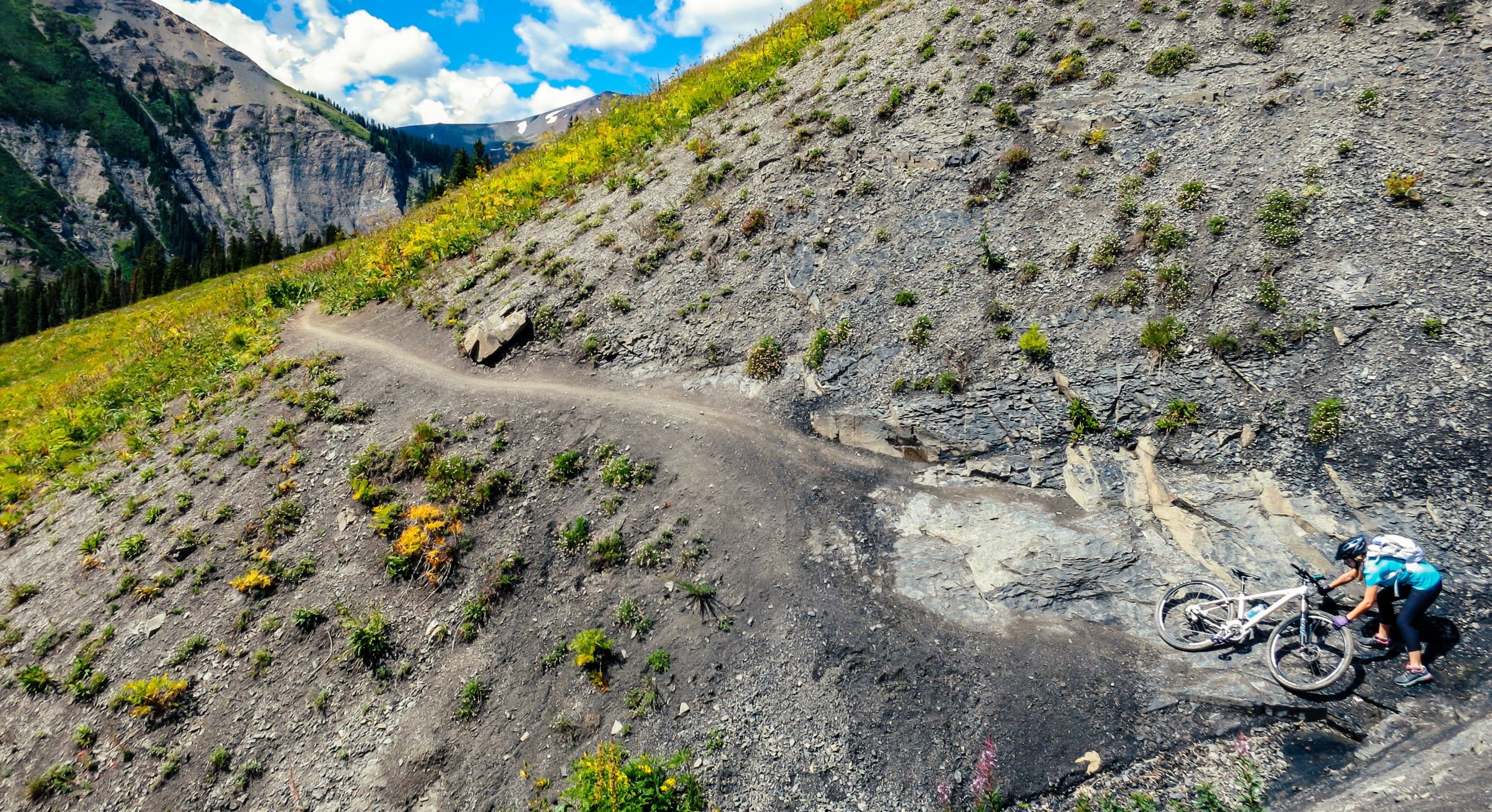 A mountain biker navigating a steep, rocky terrain while pushing their bike along a winding dirt path surrounded by lush green hills and a blue sky with scattered clouds. Trail 401 mountain bike trail.