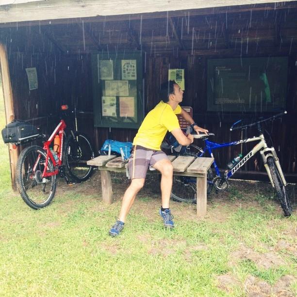 A person wearing a yellow shirt and shorts is sitting on a wooden bench under a shelter, rain falling around them. Two bicycles are parked nearby, one red and one blue. The background features informational posters. False Cape State Park (back Bay Wildlife Refuge) mountain bike trail.