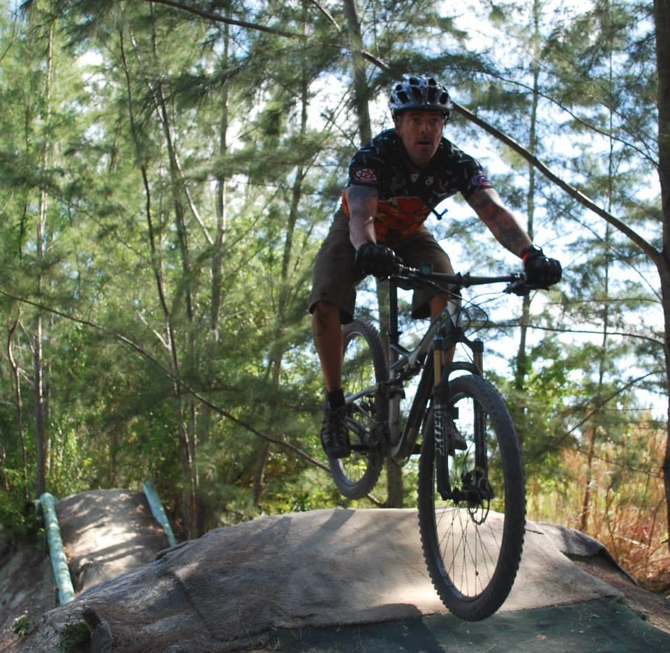 A mountain biker performing a jump on a trail surrounded by trees, showcasing dynamic action and skill. The rider is wearing a helmet, gloves, and a colorful jersey, with a rocky terrain in the foreground and a wooded background. Alafia River State Park mountain bike trail.
