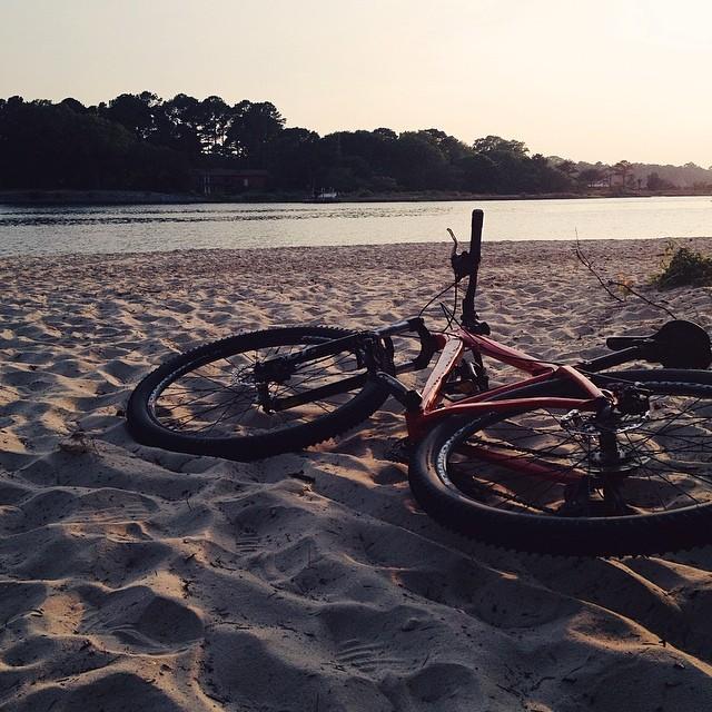 A mountain bike resting on soft sand near a calm river at sunset, surrounded by trees in the background. The warm light casts a serene atmosphere over the scene. First Landing State Park mountain bike trail.