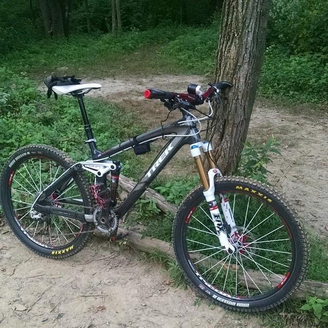 Trek Remedy 9.9: A mountain bike leaning against a tree in a forested area. The bike features a black frame with red and white accents, dual suspension, and large tires suitable for trail riding. The background shows a dirt path surrounded by greenery.