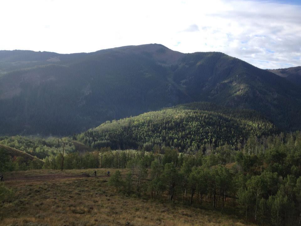 A panoramic view of a mountainous landscape featuring rolling hills and dense forests. The scene is illuminated by soft sunlight with a partly cloudy sky, highlighting the lush greenery of the trees below and the rugged terrain of the mountains in the background. Walrod Gulch / #412 mountain bike trail.