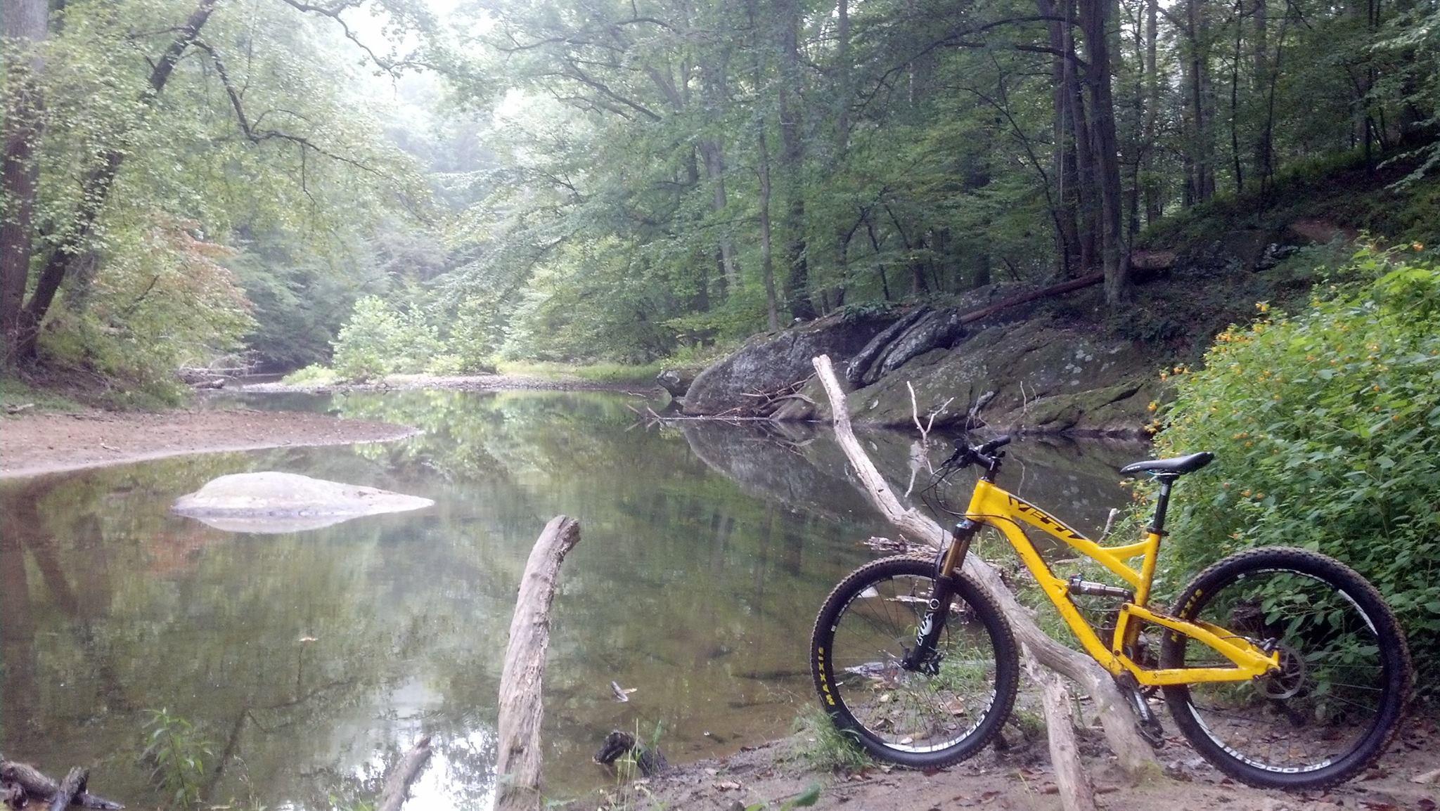 A yellow mountain bike resting beside a tranquil stream in a lush wooded area, with reflections of trees in the water and a rocky shoreline. Gunpowder Falls State Park mountain bike trail.