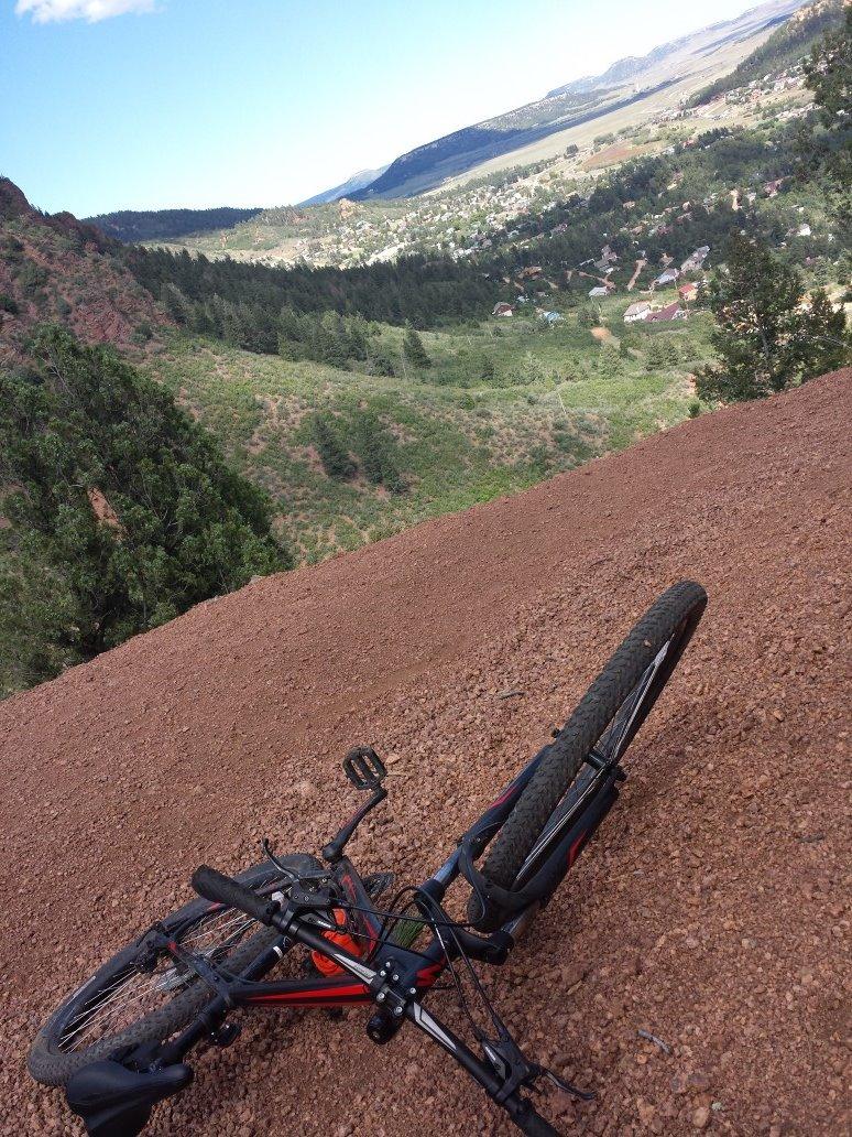 Specialized Hardrock Disc 29: A mountain bike lying on the rocky terrain of a hillside, with a panoramic view of a lush valley and distant mountains in the background under a clear blue sky.