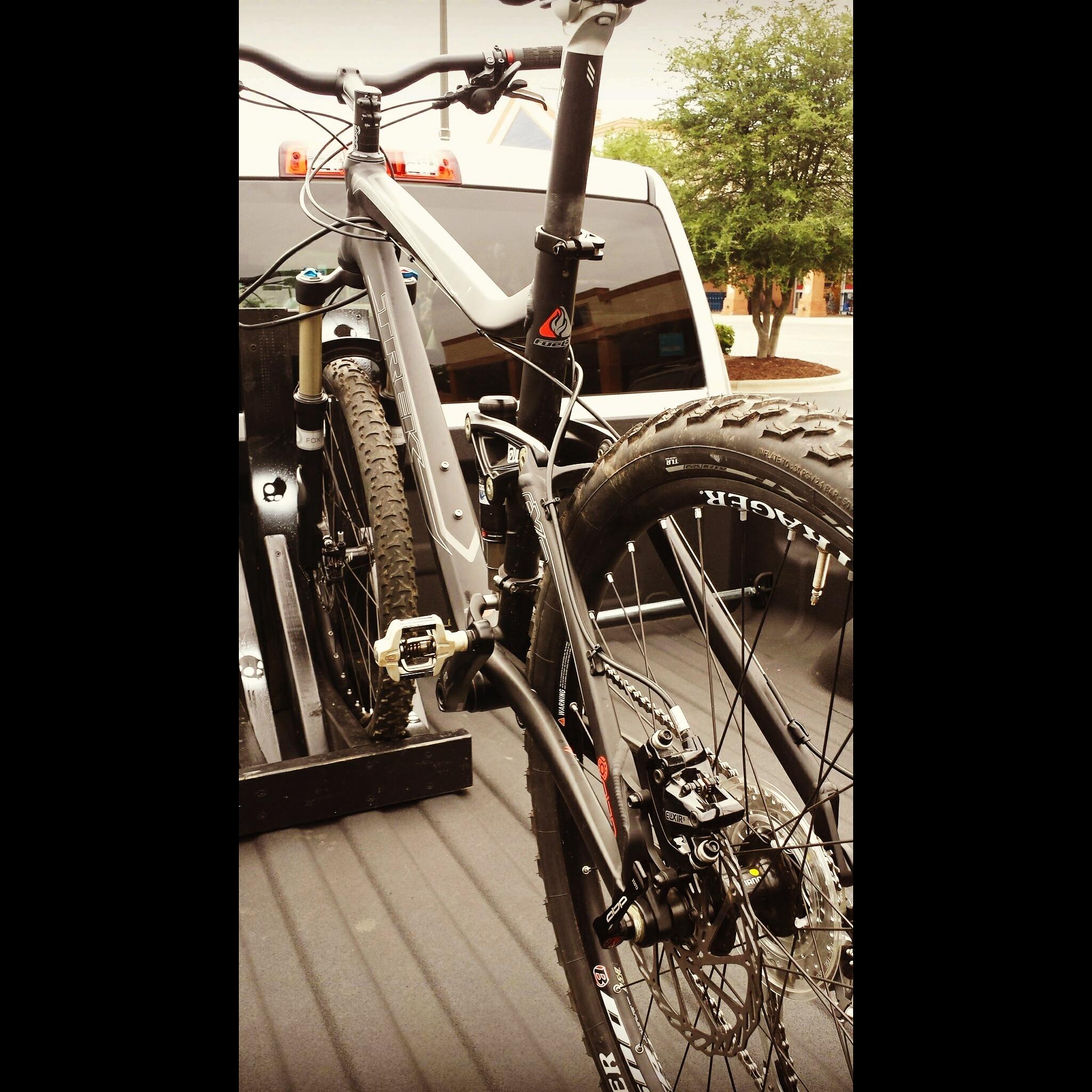 Trek Fuel EX 8: A black mountain bike secured in the bed of a pickup truck, showcasing its frame, handlebars, and large knobby tires. The background features the rear window of the truck and some greenery.