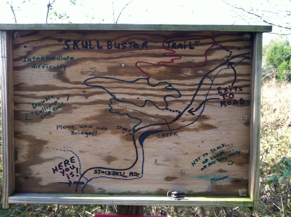 A wooden trail map sign for the "Skull Buster Trail," indicating an intermediate difficulty level. The map features a winding path with labeled points, including "Start," "Creek," and "Exits to Road." Additional notes highlight advice to walk over bridges and mention a double loop. The map includes colorful illustrations and informal annotations, and is set against a natural background. Skullbuster mountain bike trail.