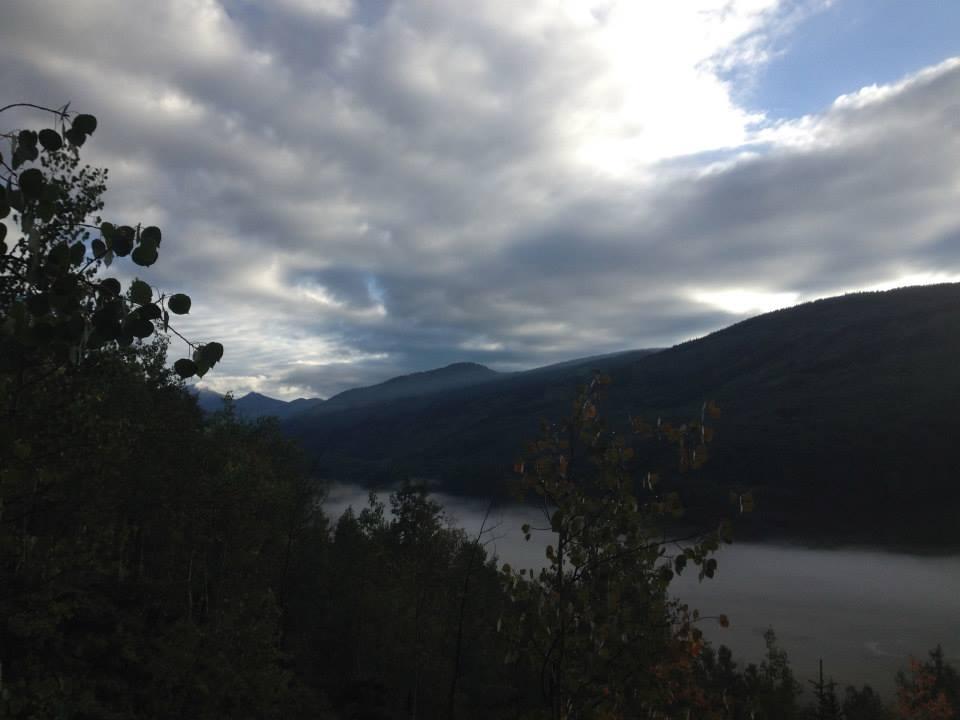 A scenic view of mountains partially covered by clouds, with a river winding through the valley below. The foreground features leafy trees, and the sky is filled with a gradient of gray and blue hues, suggesting an early morning or late afternoon setting. Warm Springs Trail / #406 mountain bike trail.