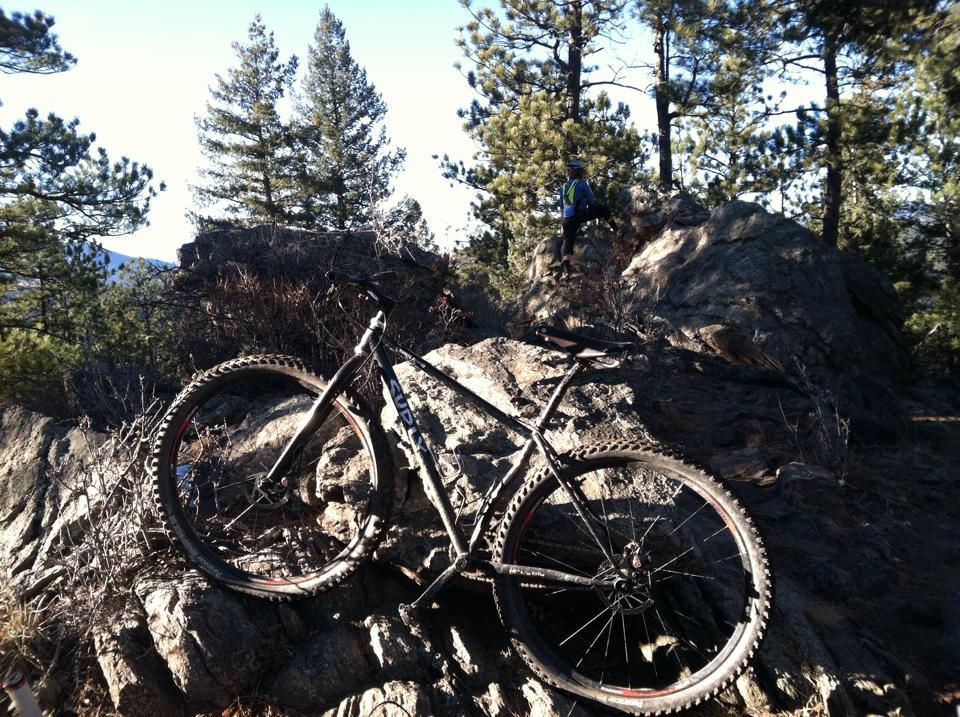 Surly Karate Monkey: A mountain bike resting on rocky terrain with pine trees in the background. A person is climbing up a large boulder in the distance. The scene captures a sunny outdoor setting, ideal for mountain biking and hiking.