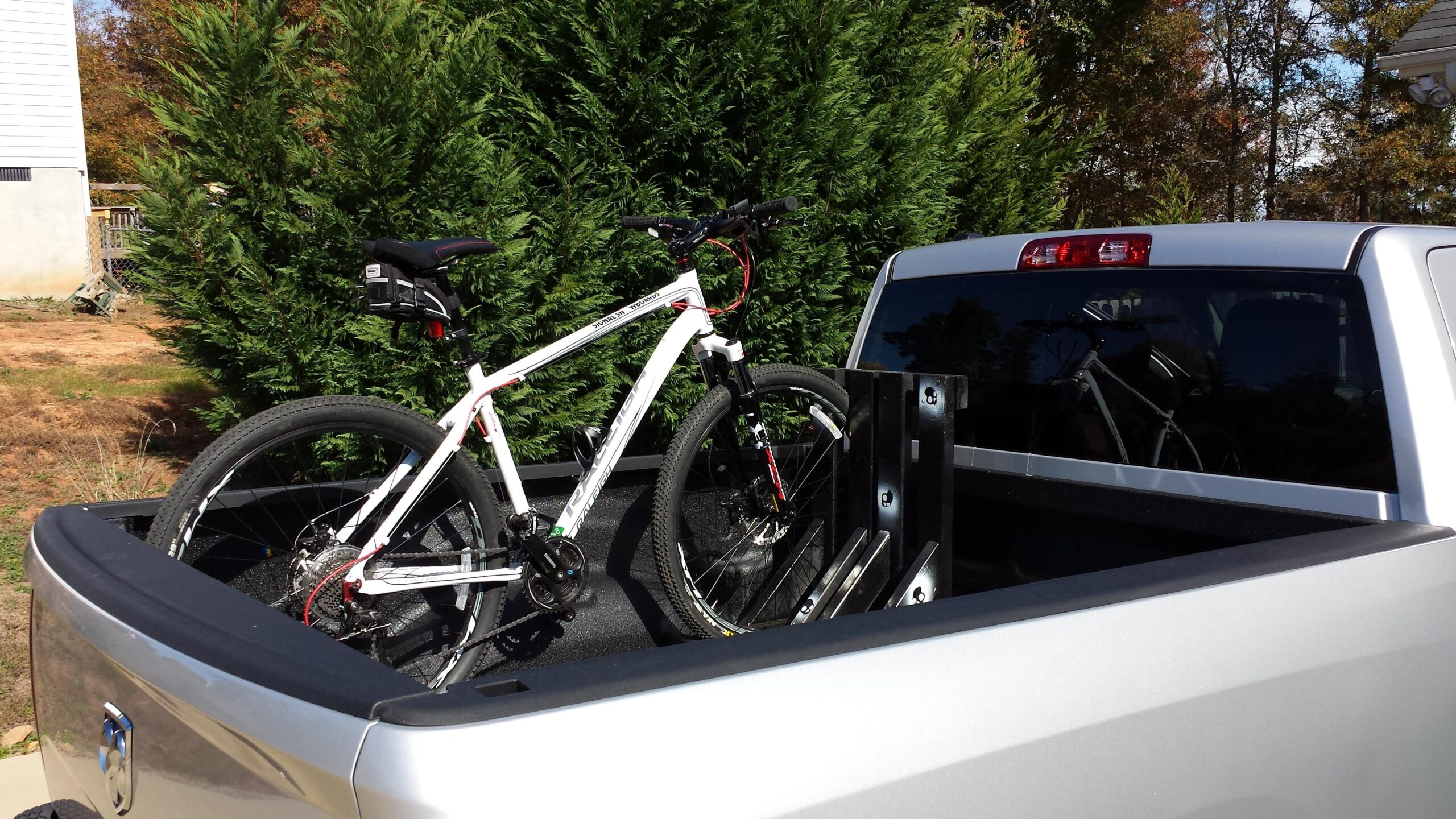 Raleigh Talus 29er: A white mountain bike is secured in the bed of a silver pickup truck, with a black bike rack visible in the background. Lush green shrubbery surrounds the scene, and a portion of a house can be seen in the background, indicating an outdoor setting.