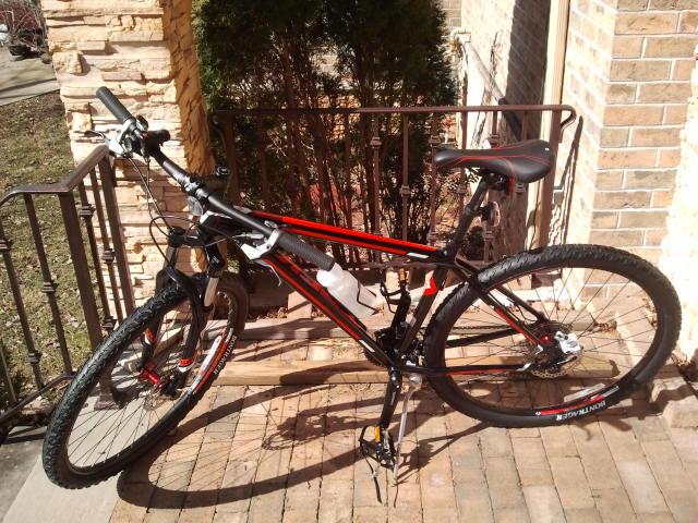 Trek Marlin: A black and red mountain bike is leaning against a railing on a brick pathway in front of a house. The bike features thick tires and a water bottle attached to the frame, with natural greenery visible in the background.