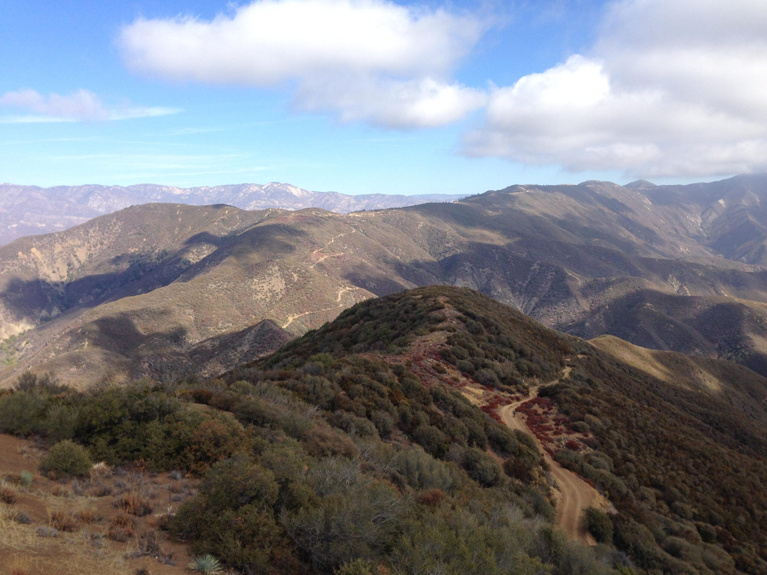 A panoramic view of rolling hills and mountains under a partly cloudy sky. The landscape is covered in green and brown vegetation, with a dirt path winding through the terrain. Sunlight casts shadows on the slopes, creating variations in color across the hills. Gridley mountain bike trail.